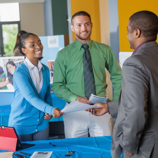 Picture of a man and a woman both holing papers and handing them to the third man. All people in the picture are dresses professionally.  