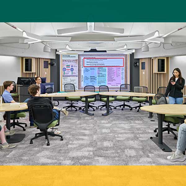 A student standing and presenting to seated students in a State Hall classroom with a green border overlay at the top and a gold border overlay at the bottom