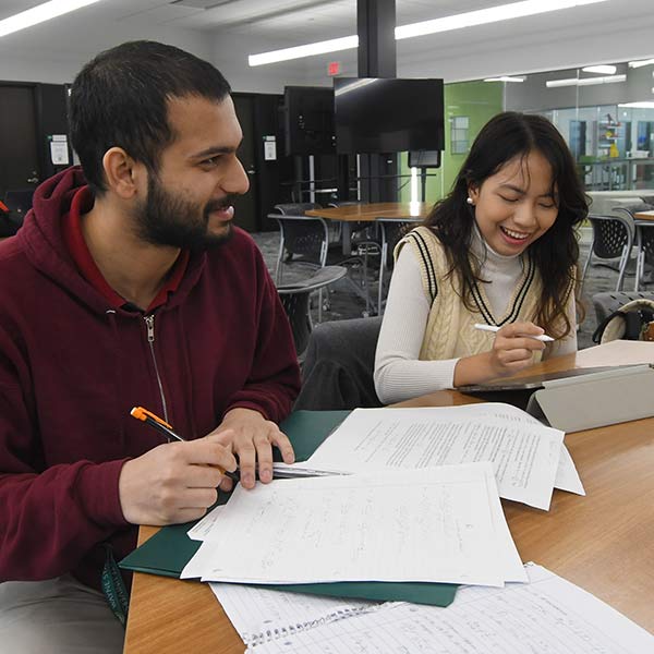 two students working at a desk together, one is working on an tablet and the other is writing on papers