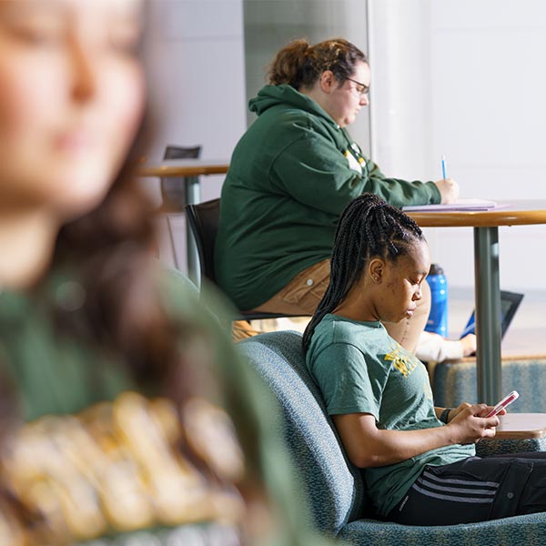 Three students working: a student in the foreground, a student sitting in chair using a cell phone, and a student sitting at a high table writing on a notepad.