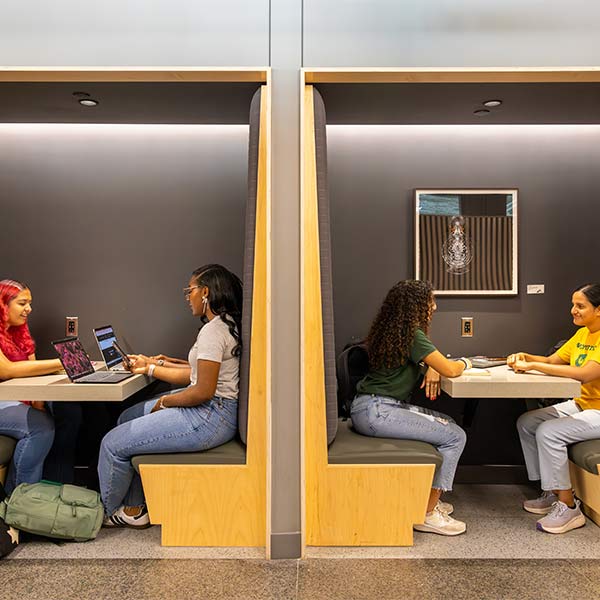 A profile view of two groups of two students sitting in hallway booths talking and working on laptops