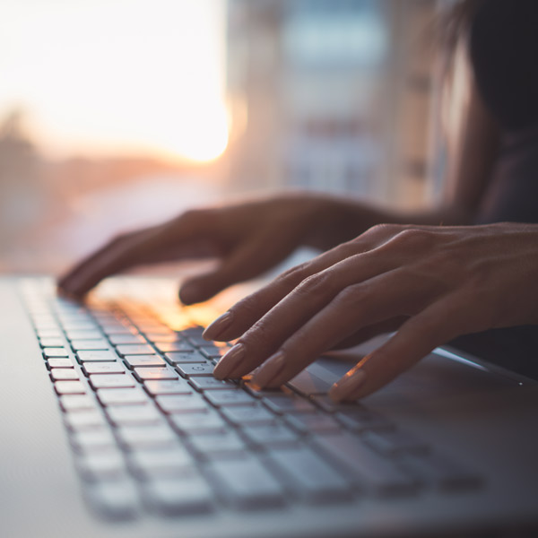 Close-up of a person's hands typing on a laptop keyboard with a soft-focus background illuminated by the warm glow of a sunset