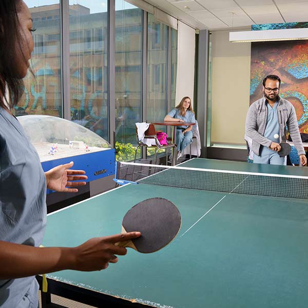 Medical students playing ping-pong in a recreation room with other students sitting at tables in the background