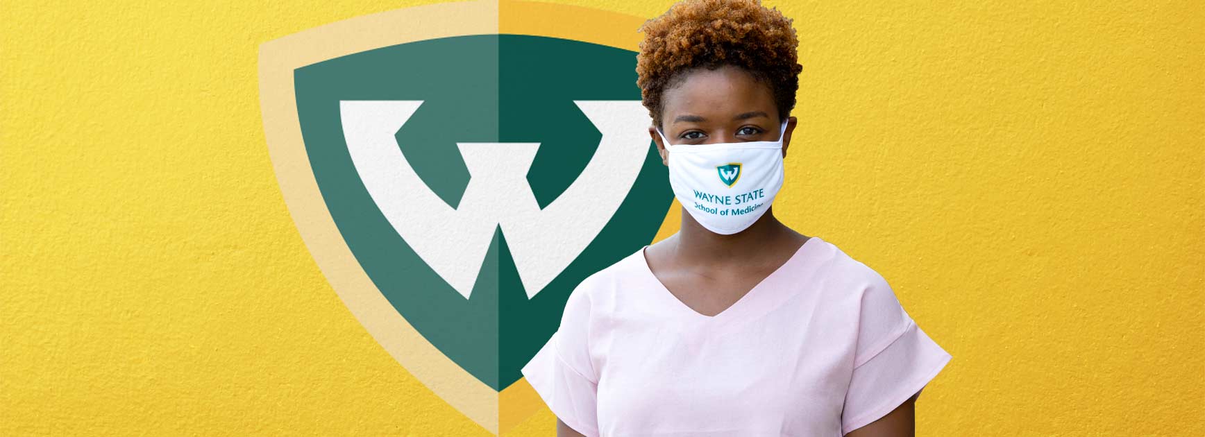 Woman wearing a cloth mask standing in front of the WSU shield on a yellow wall